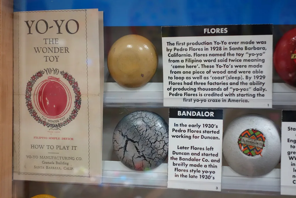 Museum case with a "Yo-Yo, The Wonder Toy" flyer from the Yo-Yo Manufacturing Co. of Santa Barbara beside Flores and Bandalor yo-yos and exhibit labels.